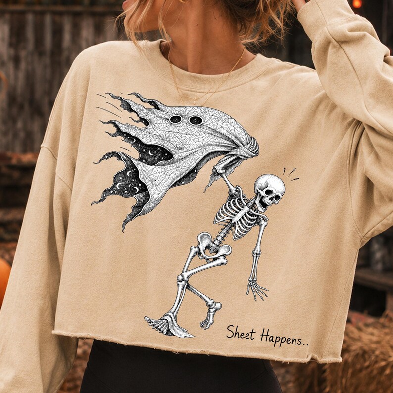 K&ouml;nnte beinhalten: Ein beiges Langarm-Sweatshirt mit offenem Saum und einem schwarz-wei&szlig;en Grafikdruck eines Skeletts, das ein Geisterlaken h&auml;lt. Das Laken zeigt ein Spinnennetz-Muster und einen Sternenhimmel im Inneren. Der Text Sheet Happens ist aufgedruckt.