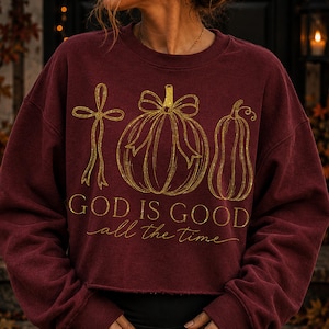 K&ouml;nnte beinhalten: Ein bordeauxrotes Langarm-Sweatshirt mit Rundhalsausschnitt und einem goldenen Grafikdruck, der eine Schleife, einen K&uuml;rbis mit Schleife und einen Zierk&uuml;rbis zeigt. Der Text "GOD IS GOOD all the time" steht in goldener Schrift unter den Motiven.