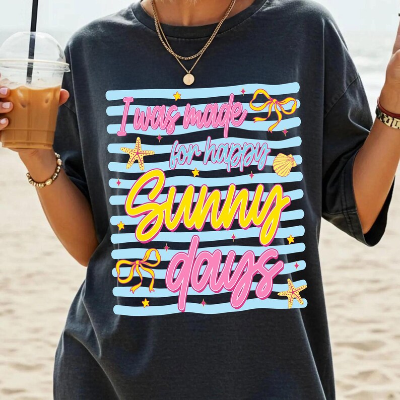 Pu&ograve; includere: T-shirt grigio scuro con la scritta "I was made for happy sunny days" in rosa e giallo. Il design include strisce blu, fiocchi, stelle marine e una conchiglia. Una t-shirt grafica casual a tema spiaggia.
