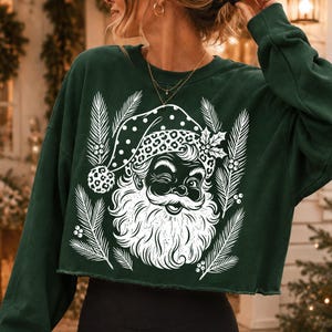Puede incluir: Una sudadera corta de manga larga en color verde oscuro con un estampado gr&aacute;fico blanco de un Santa Claus gui&ntilde;ando un ojo, con un gorro de estampado de leopardo y lunares, rodeado de ramas de pino y bayas de acebo estilizadas en el frente.