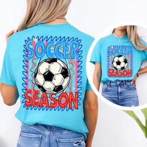 Puede incluir: Camiseta azul claro con un dise&ntilde;o de bal&oacute;n de f&uacute;tbol. El dise&ntilde;o incluye las palabras "SOCCER SEASON" en un dise&ntilde;o colorido. La camiseta es de manga corta y est&aacute; hecha de un material suave.