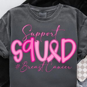 May include: A dark grey t-shirt featuring the words "Support Squad" in pink script and block letters, with a pink ribbon graphic. The phrase "#Breast Cancer" is below in pink.