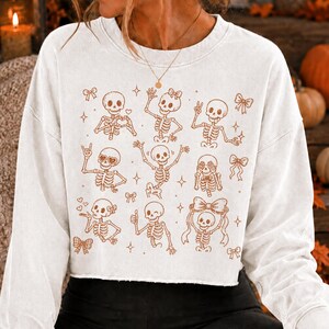 Op de afbeelding: Een wit sweatshirt met lange mouwen en ronde hals, voorzien van een bruine grafische print van meerdere skeletten in diverse houdingen, waaronder zwaaiende, hartvormige handen en vredestekens, afgewisseld met kleine sterren en strikjes.