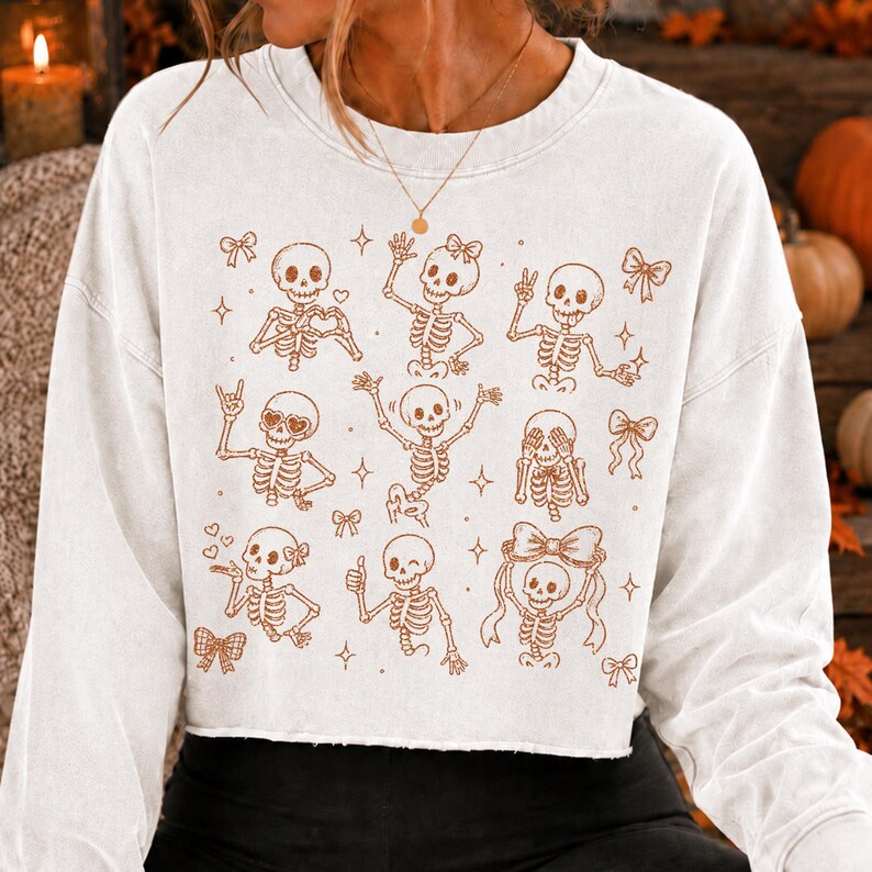 Op de afbeelding: Een wit sweatshirt met lange mouwen en ronde hals, voorzien van een bruine grafische print van meerdere skeletten in diverse houdingen, waaronder zwaaiende, hartvormige handen en vredestekens, afgewisseld met kleine sterren en strikjes.