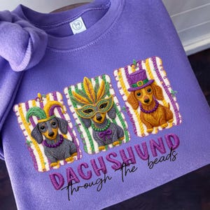 Puede incluir: Una sudadera lila con bordados de perros salchicha con disfraces de Mardi Gras. Cada perro lleva un accesorio festivo diferente: un gorro de buf&oacute;n, una m&aacute;scara de plumas y un sombrero de copa. El texto "DACHSHUND Through The beads" est&aacute; bordado debajo.