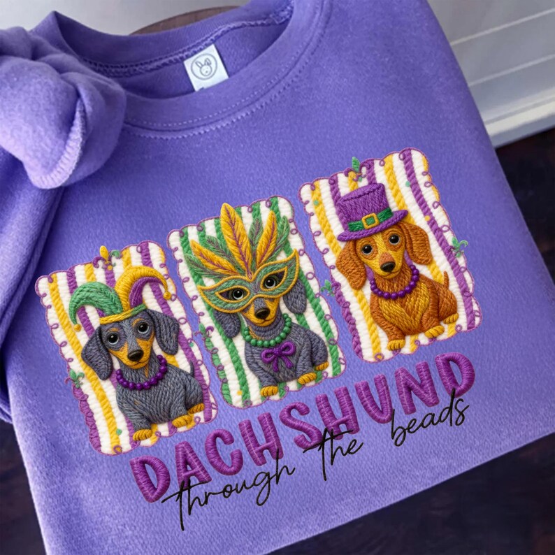 Puede incluir: Una sudadera lila con bordados de perros salchicha con disfraces de Mardi Gras. Cada perro lleva un accesorio festivo diferente: un gorro de buf&oacute;n, una m&aacute;scara de plumas y un sombrero de copa. El texto "DACHSHUND Through The beads" est&aacute; bordado debajo.