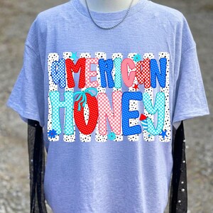 May include: A light heather gray short-sleeve t-shirt featuring a graphic print with the text AMERICAN HONEY in large, multi-colored, patterned bubble letters. The design includes red, blue, and pink polka dots, checkers, and small star accents.
