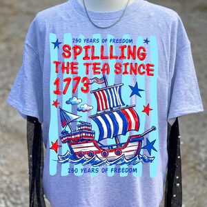 Pu&ograve; includere: T-shirt grigio chiaro con un design patriottico. La grafica presenta una nave con vele rosse, bianche e blu e il testo "Spilling the Tea Since 1773" e "250 Years of Freedom".