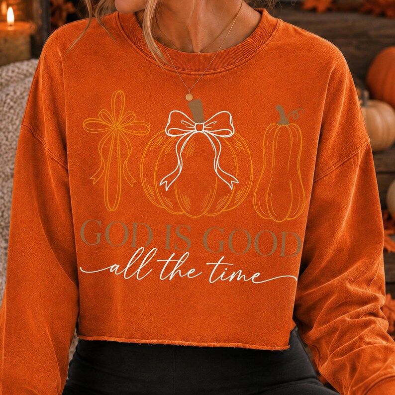 Puede incluir: Una sudadera naranja de manga larga con un estampado gr&aacute;fico de una cruz, una calabaza con un lazo blanco y una calabaza alargada. El texto "GOD IS GOOD" est&aacute; impreso en letras marrones sobre el texto en cursiva blanca "all the time".