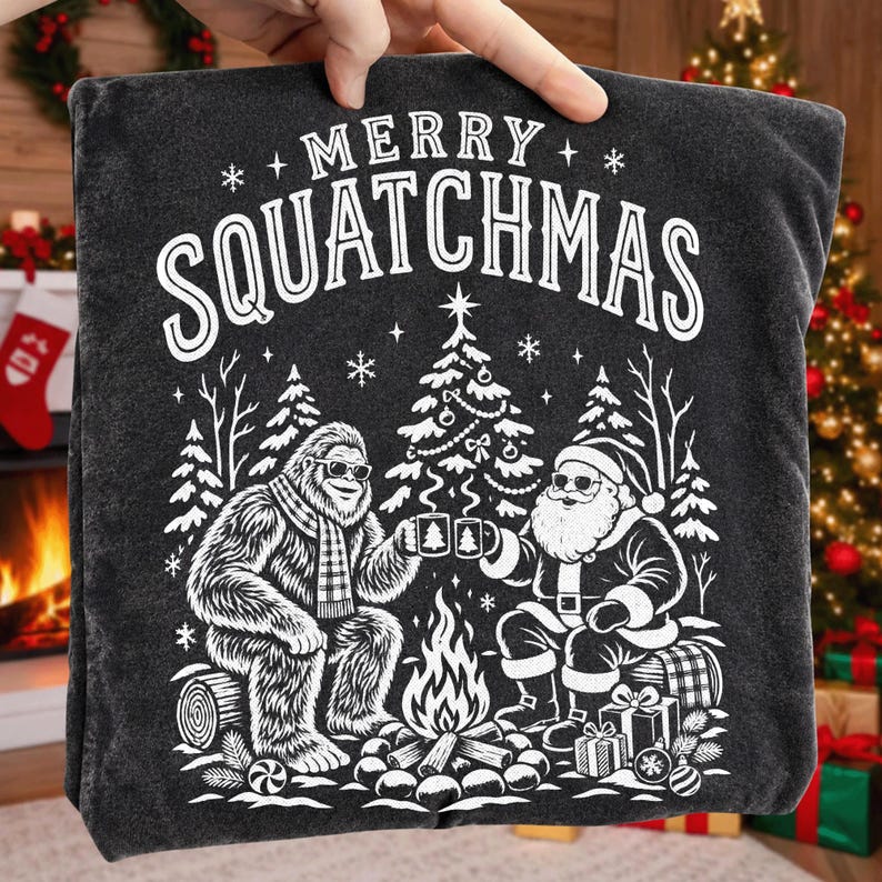 K&ouml;nnte beinhalten: Ein anthrazitfarbenes T-Shirt mit einem wei&szlig;en Grafikdruck und dem Text MERRY SQUATCHMAS. Das Motiv zeigt einen Sasquatch und den Weihnachtsmann mit Sonnenbrillen, die an einem Lagerfeuer vor einem geschm&uuml;ckten Weihnachtsbaum sitzen.