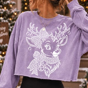 May include: A light purple long-sleeve cropped sweatshirt featuring a white graphic print of a reindeer with large eyes, wearing a leopard print bow and scarf, with ornaments hanging from its antlers. The fabric has a washed, textured appearance.
