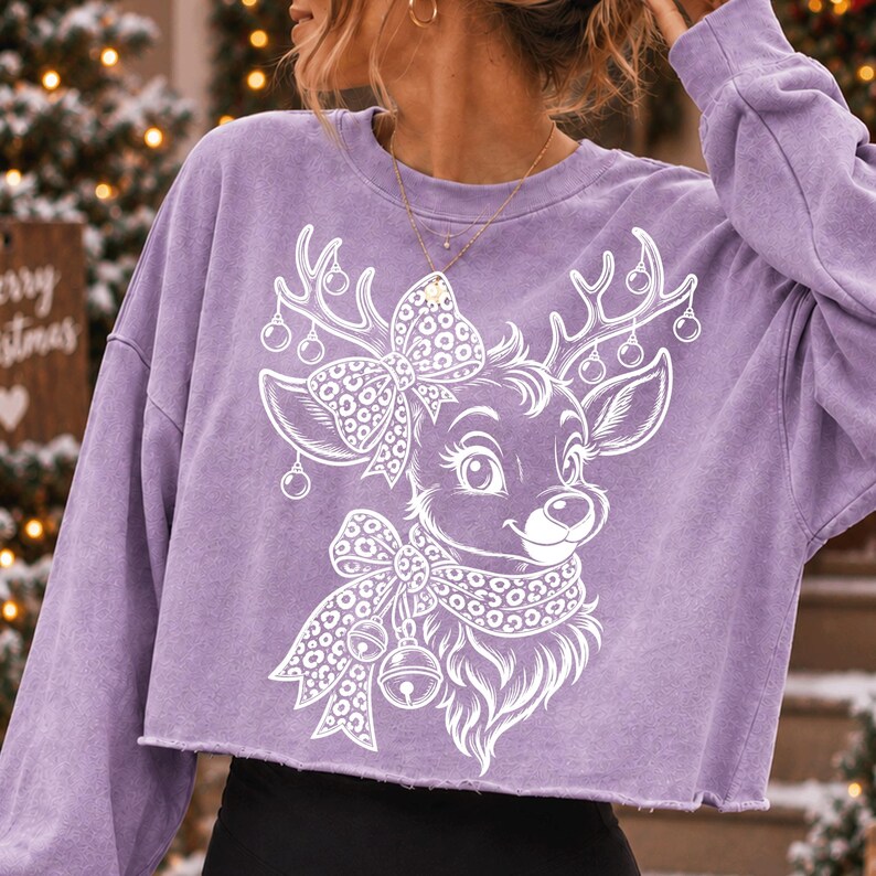 May include: A light purple long-sleeve cropped sweatshirt featuring a white graphic print of a reindeer with large eyes, wearing a leopard print bow and scarf, with ornaments hanging from its antlers. The fabric has a washed, textured appearance.