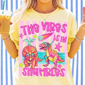 Peut inclure: T-shirt jaune clair avec un motif &agrave; rayures roses et blanches. Le motif pr&eacute;sente le texte "THE VIBES IS IN SHAMBLES" avec des dinosaures de dessins anim&eacute;s, des &eacute;toiles et une com&egrave;te. Les dinosaures portent des lunettes de soleil et un tuba.