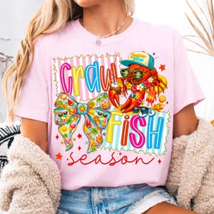 May include: Pale pink t-shirt with a vibrant graphic. It shows a cartoon lobster in sunglasses and a cap, with the words "Crawfish Season". The design includes a bow and tropical motifs.