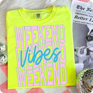 K&ouml;nnte beinhalten: Ein neongelbes T-Shirt mit den Worten "WEEKEND VIBES WEEKEND" in rosa und blauer Schrift. Das Shirt ist von der Marke Comfort Colors. Das Design ist mittig auf der Vorderseite. Das Shirt wird auf einer rosa-wei&szlig; gestreiften Oberfl&auml;che pr&auml;sentiert.