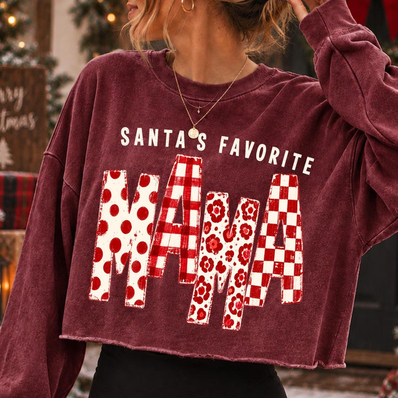 K&ouml;nnte beinhalten: Ein bordeauxrotes, lang&auml;rmeliges, bauchfreies Sweatshirt mit dem wei&szlig;en Schriftzug "SANTA'S FAVORITE" &uuml;ber dem Wort "MAMA". Die Buchstaben in "MAMA" sind mit verschiedenen rot-wei&szlig;en Mustern wie Punkten, Karos, Blumen und Schachbrettmustern gef&uuml;llt.