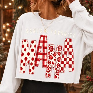 K&ouml;nnte beinhalten: Ein wei&szlig;es, lang&auml;rmeliges, bauchfreies Sweatshirt mit dem Schriftzug SANTA'S FAVORITE MAMA in roten und wei&szlig;en Mustern. Die Buchstaben MAMA sind mit roten Punkten, einem roten Karomuster, roten Blumenmustern und einem roten Schachbrettmuster gef&uuml;llt.