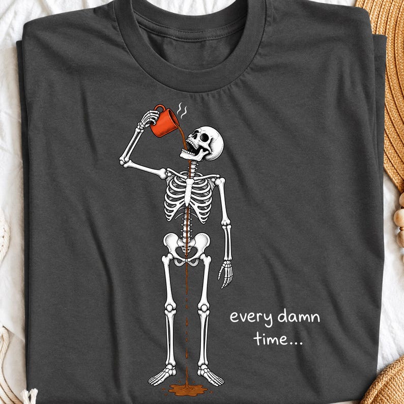 May include: A dark grey t-shirt featuring a white skeleton graphic drinking from an orange mug. The coffee flows through the skeleton's ribcage and down to a puddle on the floor. The text "every damn time..." is printed in white on the right side.