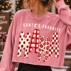K&ouml;nnte beinhalten: Ein altrosa lang&auml;rmeliges, bauchfreies Sweatshirt mit dem wei&szlig;en Schriftzug SANTA'S FAVORITE &uuml;ber dem Wort MAMA. Die Buchstaben von MAMA sind mit verschiedenen rot-wei&szlig;en Mustern gef&uuml;llt, darunter Punkte, Vichy-Karo und Blumenmuster.