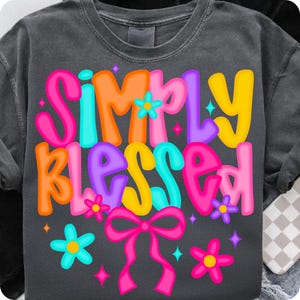 Puede incluir: Sudadera gris oscuro con la frase "SIMPLY BLESSED" en letras coloridas. El diseño incluye flores, lazos y estrellas en tonos rosa, naranja, amarillo, azul y morado.