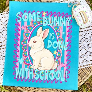 Może przedstawiać: Turkusowa tkanina z napisem "SOME BUNNY IS DONE WITH SCHOOL!" w kolorze jasnoniebieskim, r&oacute;żowym i złotym. Biały kr&oacute;lik znajduje się w owalnej ramce. Projekt zawiera gwiazdy i dekoracyjne linie.