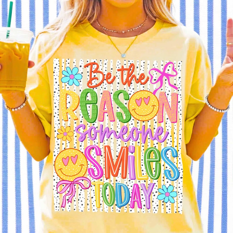 May include: Yellow t-shirt with the phrase "Be the Reason Someone Smiles Today" in colorful, playful lettering. The design includes smiley faces, flowers, and bows. The shirt has short sleeves.