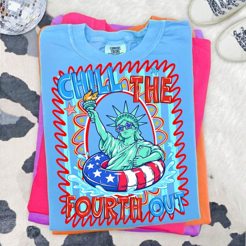 May include: A light blue t-shirt with a graphic of the Statue of Liberty wearing star-shaped sunglasses and a patriotic inner tube. The text "CHILL THE FOURTH OUT" is displayed in red and blue. The shirt is folded on top of other colorful shirts.