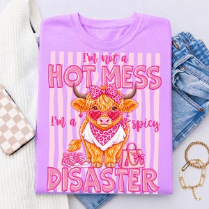 K&ouml;nnte beinhalten: Lilafarbenes T-Shirt mit einer Grafik einer Highland-Kuh mit herzf&ouml;rmiger Sonnenbrille, Schleife und Halstuch. Der Text lautet: "I'm not a HOT MESS, I'm a spicy DISASTER."