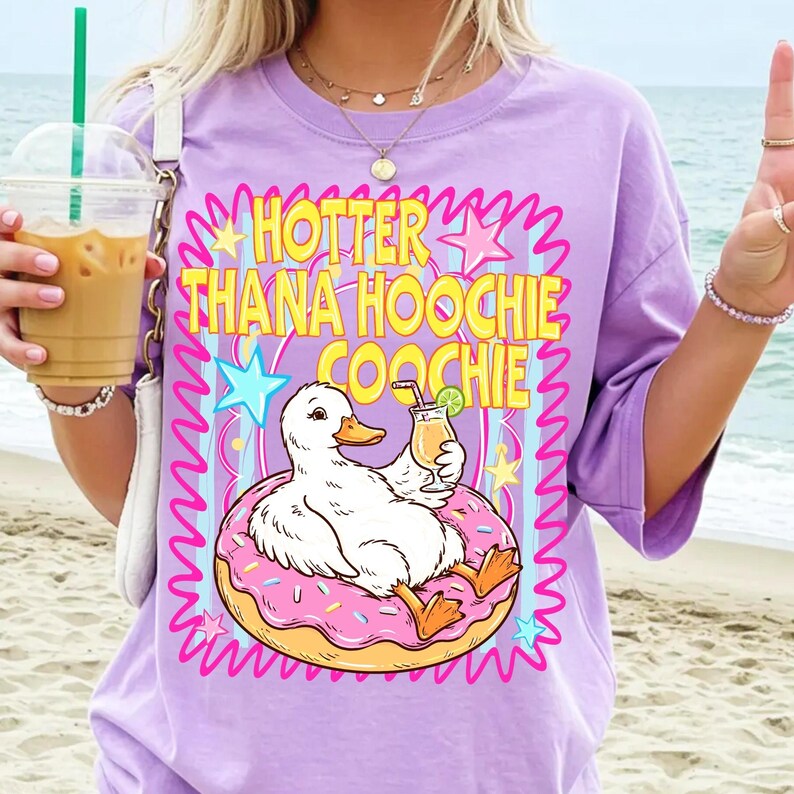 Może przedstawiać: Liliowa koszulka z grafiką białej kaczki na r&oacute;żowym pączku, trzymającej koktajl. Tekst "HOTTER THANA HOOCHIE COOCHIE" jest w kolorze ż&oacute;łtym i r&oacute;żowym. Koszulka ma kr&oacute;tkie rękawy. Obraz ma tło plażowe.