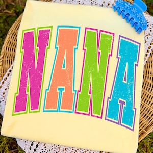 May include: A light yellow t-shirt with the word "NANA" printed in large, distressed, multi-colored letters. The letters are outlined in a contrasting color. A blue floral hair clip rests on the top right corner of the shirt.