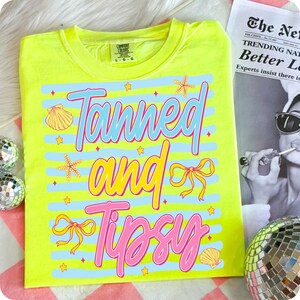 Peut inclure: Un t-shirt jaune n&eacute;on Comfort Colors avec les mots "Tanned and Tipsy" en rose, bleu et jaune. Le motif comprend des coquillages, des &eacute;toiles de mer, des n&oelig;uds et des &eacute;toiles. Le t-shirt est sur une surface ray&eacute;e rose et blanche.