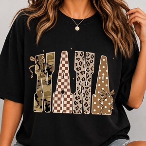 May include: A black short-sleeved t-shirt featuring the word NANA printed in large block letters. Each letter has a unique pattern: camouflage, brown and white checkerboard, leopard print, and brown with white polka dots, accented by two bows.