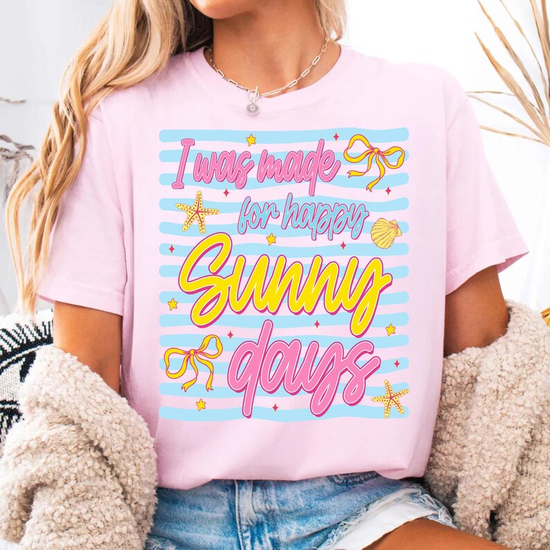 Pu&ograve; includere: T-shirt rosa chiaro con la scritta "I was made for happy sunny days" in un design colorato. Il design include stelle marine, fiocchi e una conchiglia. La maglietta ha strisce orizzontali blu.