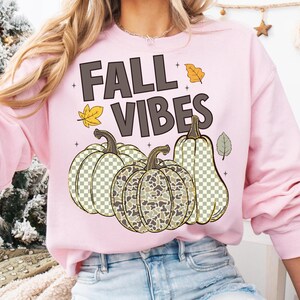 Puede incluir: Una sudadera de cuello redondo rosa claro con el texto FALL VIBES en letras marr&oacute;n oscuro. El dise&ntilde;o incluye tres calabazas con un patr&oacute;n de cuadros y camuflaje, acentuadas por peque&ntilde;as ilustraciones de hojas de oto&ntilde;o amarillas y verdes.