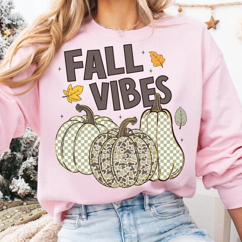 Puede incluir: Una sudadera de cuello redondo rosa claro con el texto FALL VIBES en letras marr&oacute;n oscuro. El dise&ntilde;o incluye tres calabazas con un patr&oacute;n de cuadros y camuflaje, acentuadas por peque&ntilde;as ilustraciones de hojas de oto&ntilde;o amarillas y verdes.
