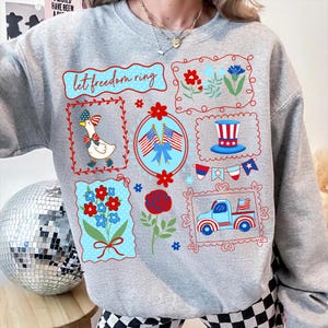 May include: A light gray sweatshirt with a patriotic design. The design includes the text "let freedom ring", a goose wearing a bow, flowers, American flags, and a truck. The graphics are in red, white, and blue.