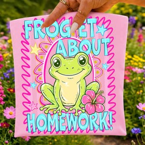 K&ouml;nnte beinhalten: Rosafarbenes T-Shirt mit einer Cartoon-Frosch-Illustration. Der Frosch ist gr&uuml;n mit gelbem Bauch und einer rosa Blume. Der Text "FROG ET ABOUT HOMEWORK!" ist blau. Das Design umfasst Sterne und einen wellenf&ouml;rmigen Rand.