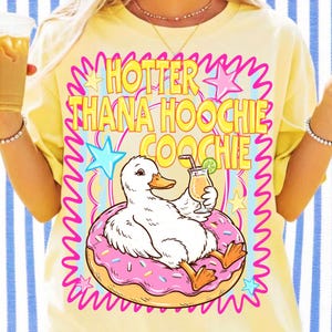 Może przedstawiać: Ż&oacute;łta koszulka z grafiką białej kaczki siedzącej na r&oacute;żowym pączku, trzymającej koktajl. Tekst "HOTTER THANA HOOCHIE COOCHIE" znajduje się nad kaczką w kolorze ż&oacute;łtym i r&oacute;żowym. Koszulka ma kr&oacute;tkie rękawy.