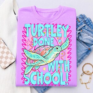 以下が含まれることがあります： ターコイズとピンクで「TURTLEY DONE WITH SCHOOL！」と書かれた薄紫色のTシャツ。ピンクとティール色の甲羅を持つ漫画のウミガメが中央にいます。シャツは白いセーター、ジーンズ、アクセサリーと一緒に背景に折りたたまれています。