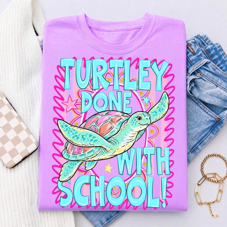 以下が含まれることがあります： ターコイズとピンクで「TURTLEY DONE WITH SCHOOL！」と書かれた薄紫色のTシャツ。ピンクとティール色の甲羅を持つ漫画のウミガメが中央にいます。シャツは白いセーター、ジーンズ、アクセサリーと一緒に背景に折りたたまれています。