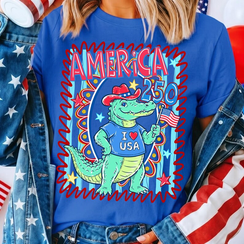Puede incluir: Una camiseta azul con un dise&ntilde;o patri&oacute;tico. La camiseta presenta la palabra "AMERICA" sobre el n&uacute;mero "250". Un cocodrilo de dibujos animados con un sombrero de vaquero y una camiseta que dice "I ❤️ USA" sostiene una bandera estadounidense.