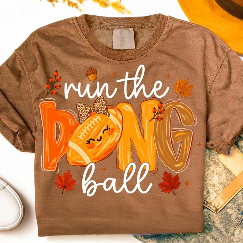 Puede incluir: Una camiseta marr&oacute;n de manga corta con el texto run the ball en letra blanca. El dise&ntilde;o incluye un bal&oacute;n de f&uacute;tbol americano con un lazo de estampado de leopardo, letras naranjas y marrones, bayas rojas y hojas de arce naranjas.