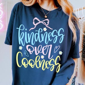 May include: Navy blue t-shirt with the words "Kindness Over Coolness" in a whimsical, colorful design. The text is in light blue, pink, and yellow, with small flower and heart accents. A pink bow is at the top.