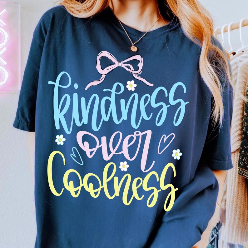 May include: Navy blue t-shirt with the words "Kindness Over Coolness" in a whimsical, colorful design. The text is in light blue, pink, and yellow, with small flower and heart accents. A pink bow is at the top.