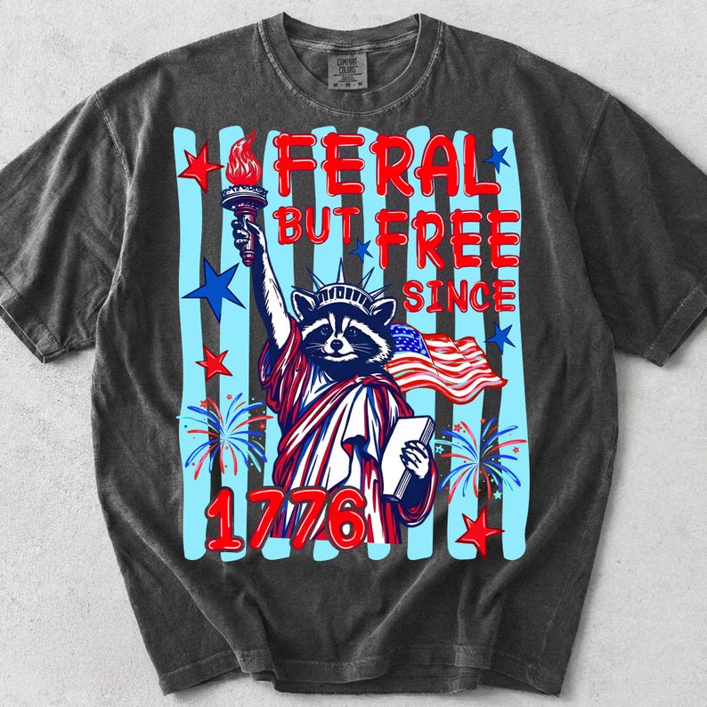 May include: Dark gray t-shirt with a graphic of a raccoon dressed as the Statue of Liberty, holding a torch and a tablet. The text reads "FERAL BUT FREE SINCE 1776" with red and blue accents. The design includes stars, fireworks, and the American flag.