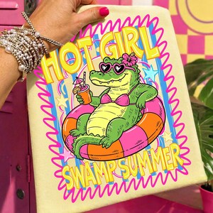 May include: A light yellow towel featuring a cartoon alligator wearing heart-shaped sunglasses and a pink bikini, floating in an orange and pink inner tube. The text "HOT GIRL SWAMP SUMMER" is displayed in yellow and pink wavy letters.