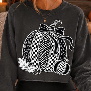 May include: A charcoal gray long-sleeve sweatshirt featuring a large white graphic print of a pumpkin decorated with checkered, wavy, and leaf patterns. A large bow sits atop the pumpkin, accompanied by a smaller pumpkin and oak leaves.