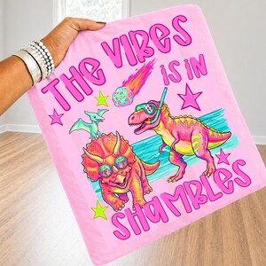 Peut inclure: Couverture rose avec le texte "The Vibes Is In Shambles" et des dinosaures de dessins anim&eacute;s portant des lunettes de soleil et un tuba. La couverture a un design color&eacute; avec des &eacute;toiles et une m&eacute;t&eacute;orite.
