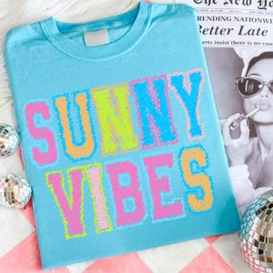 May include: A light blue t-shirt with the words "SUNNY VIBES" in large, colorful, embroidered letters. The text is outlined in pink, yellow, and orange. The shirt is folded on a pink and white surface.