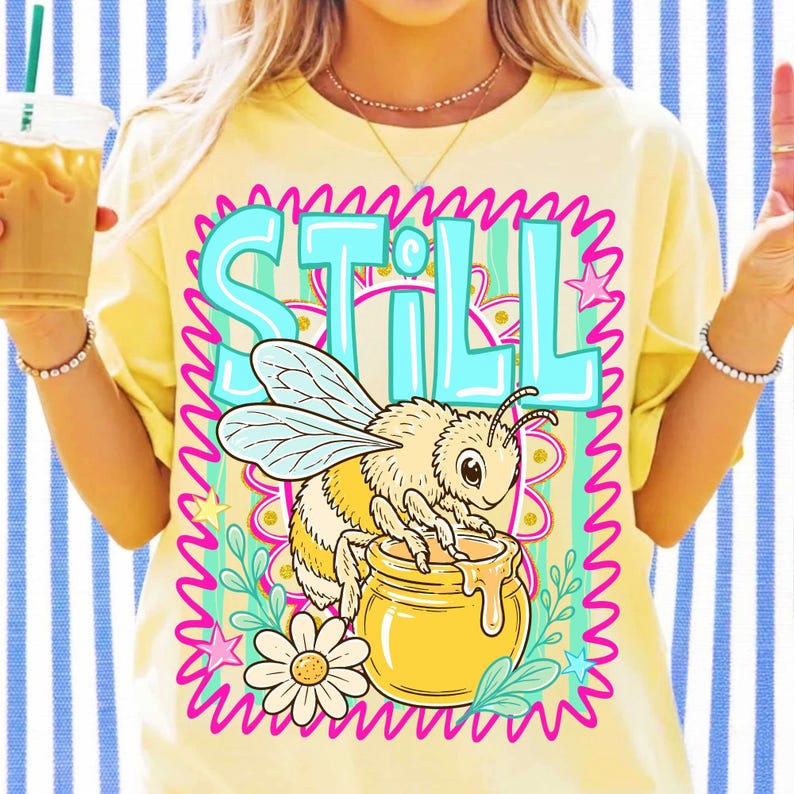 Op de afbeelding: Lichtgeel T-shirt met een afbeelding van een bij en een honingpot, met het woord "STILL" in turquoise. De bij is omgeven door een roze en groen bloemenontwerp. Het shirt heeft een lichtgele kleur.
